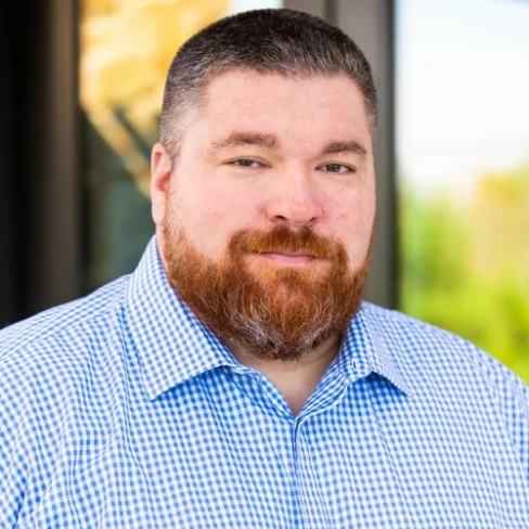 Man with short dark hair and reddish beard wearing a blue and white checkered shirt, standing outdoors with a blurred background.