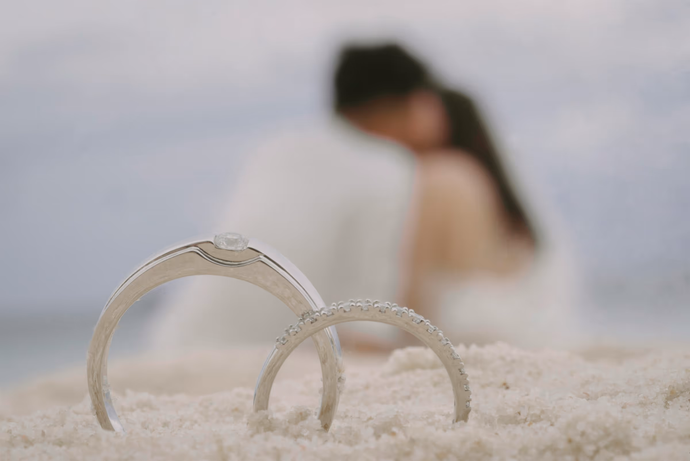 Wedding rings placed in the sand with a blurred couple embracing in the background — cinematic shot representing love, connection, and timeless wedding memories.