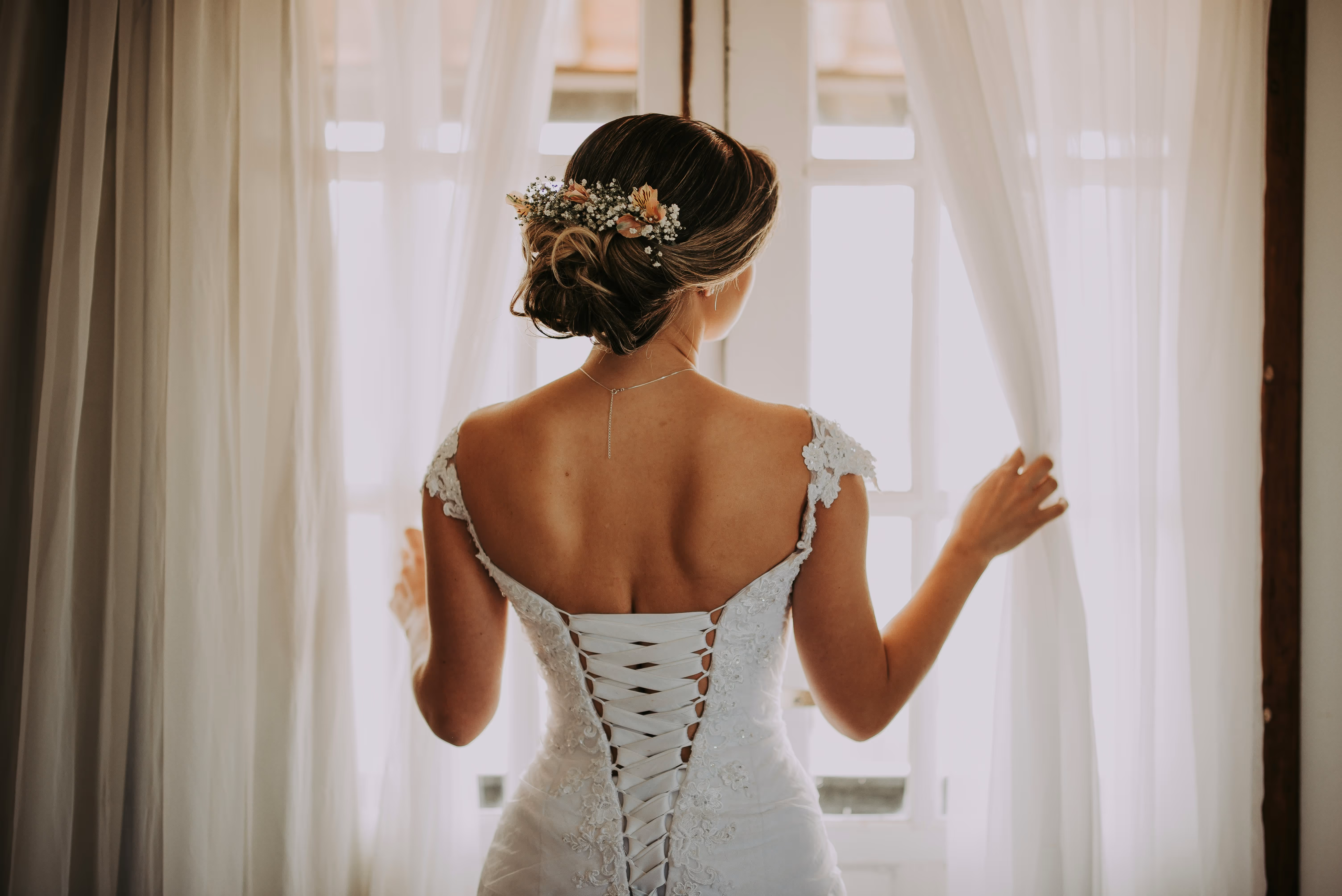 A Bride in white dress looking at a window with hair decorations and jewelry on a wedding day