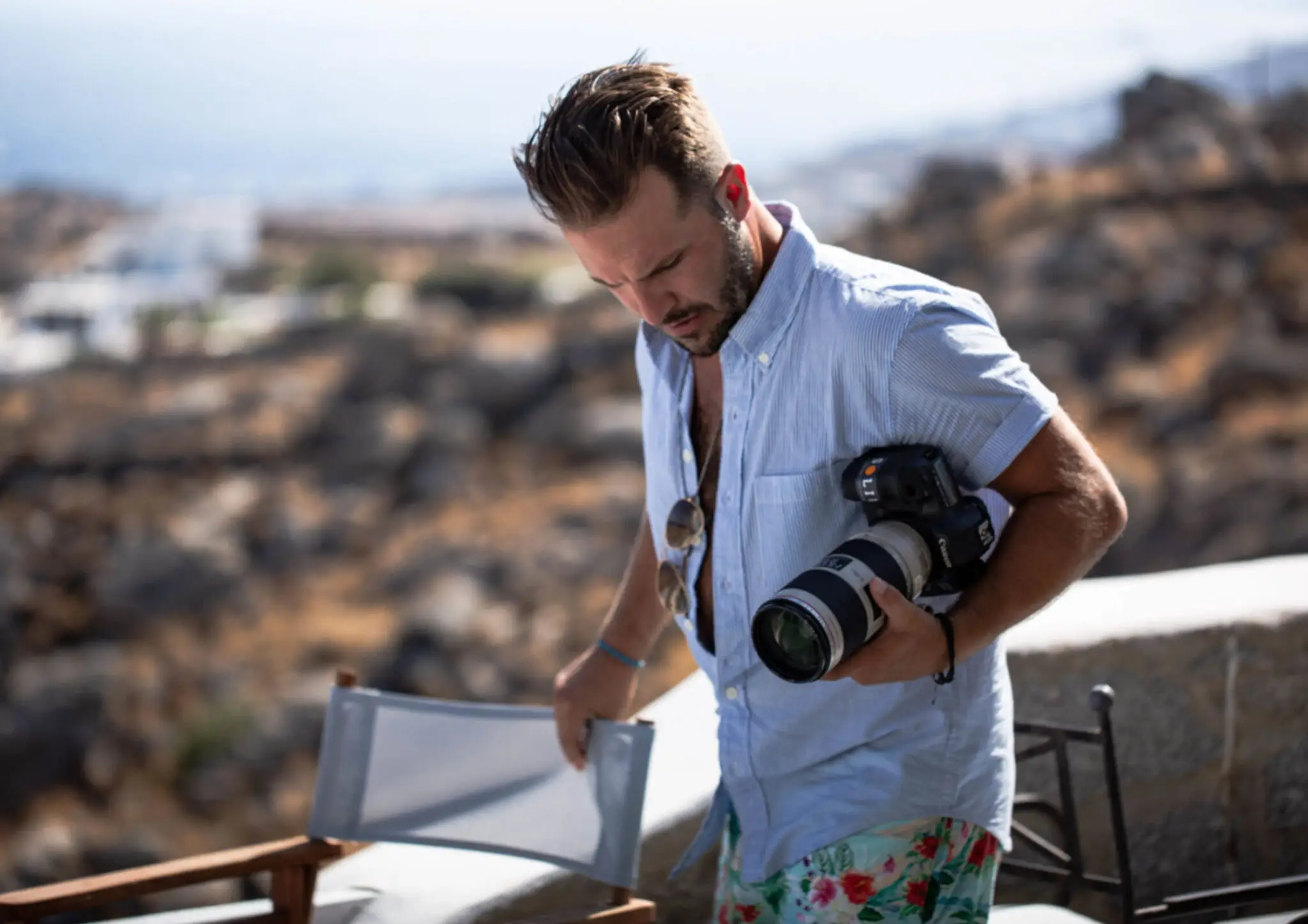 Frankie Batista with floral shorts and light blue shirt holding a professional camera outdoors with rocky hills in the background.