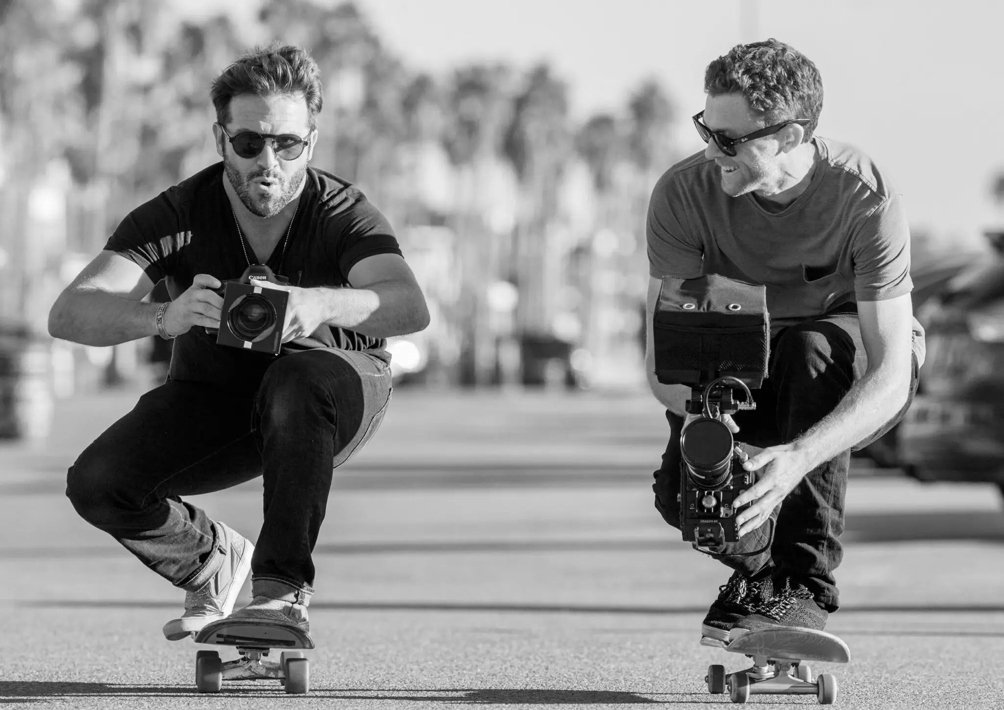 Two men crouched on skateboards filming with cameras, one holding a Canon camera and the other a professional video camera.