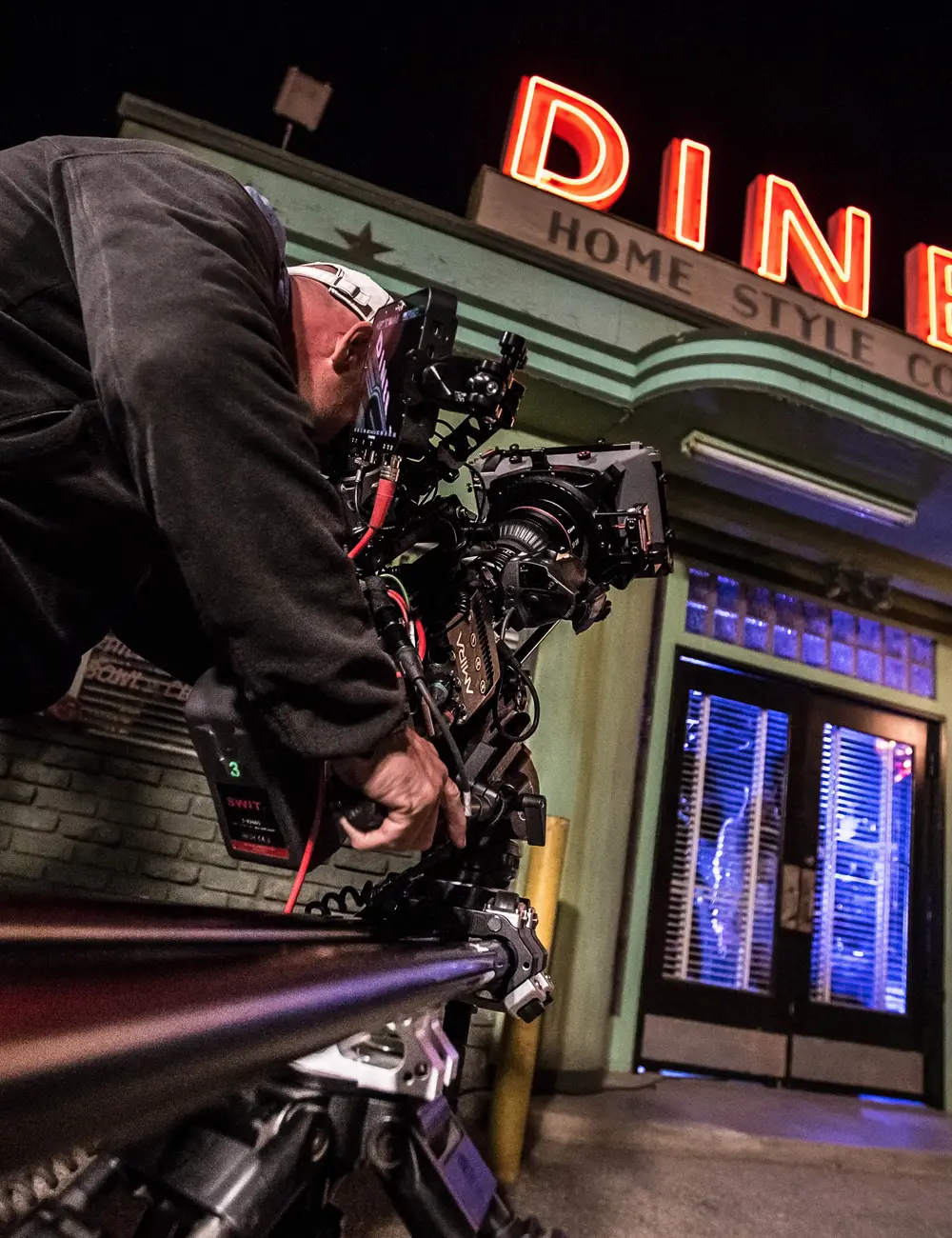 Cameraman Thomas operates a professional video camera mounted on a slider outside a diner with neon 'DINE' sign at night.