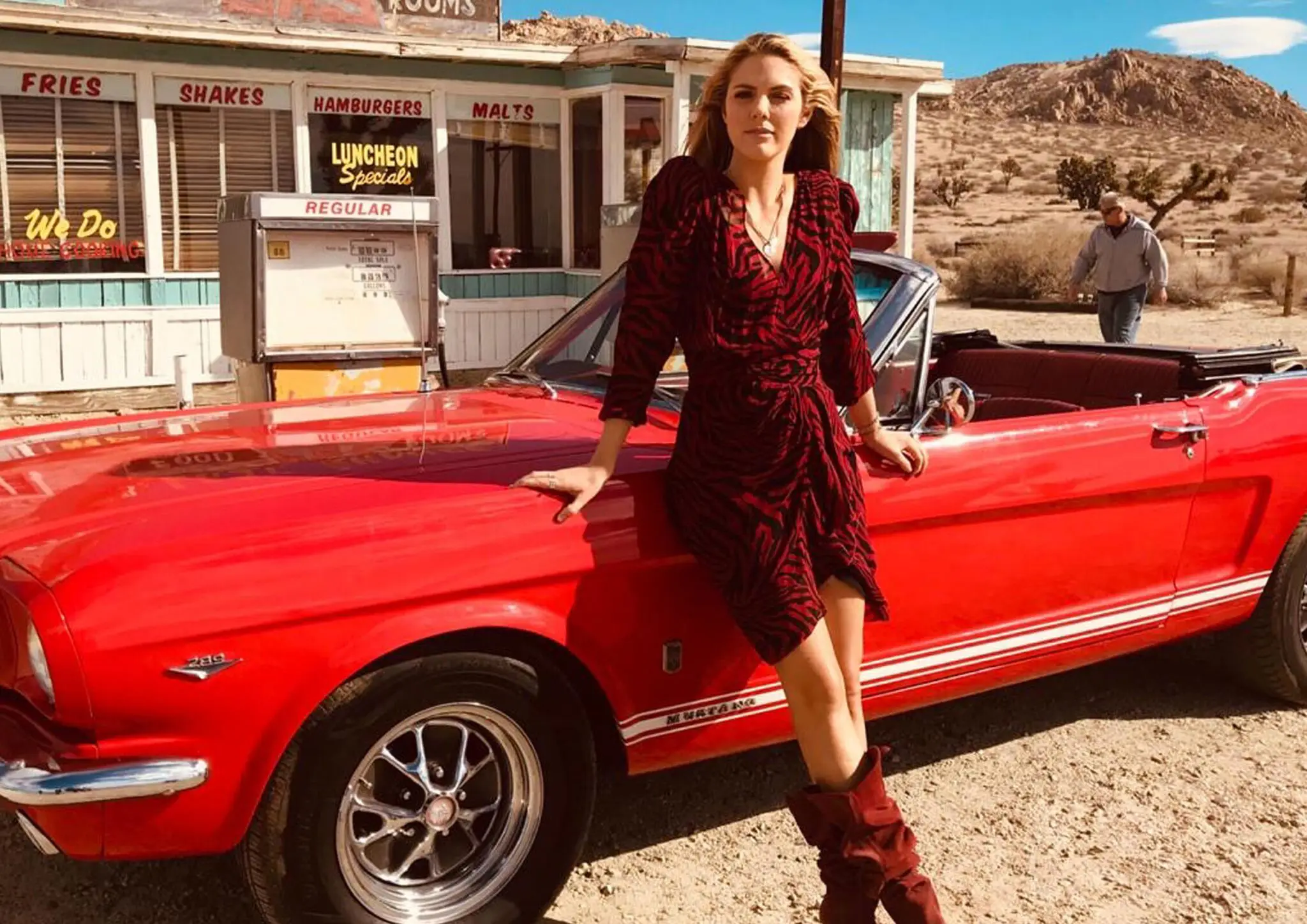 Woman in red and black dress leaning against a red vintage Ford Mustang convertible near a retro gas station in a desert landscape.