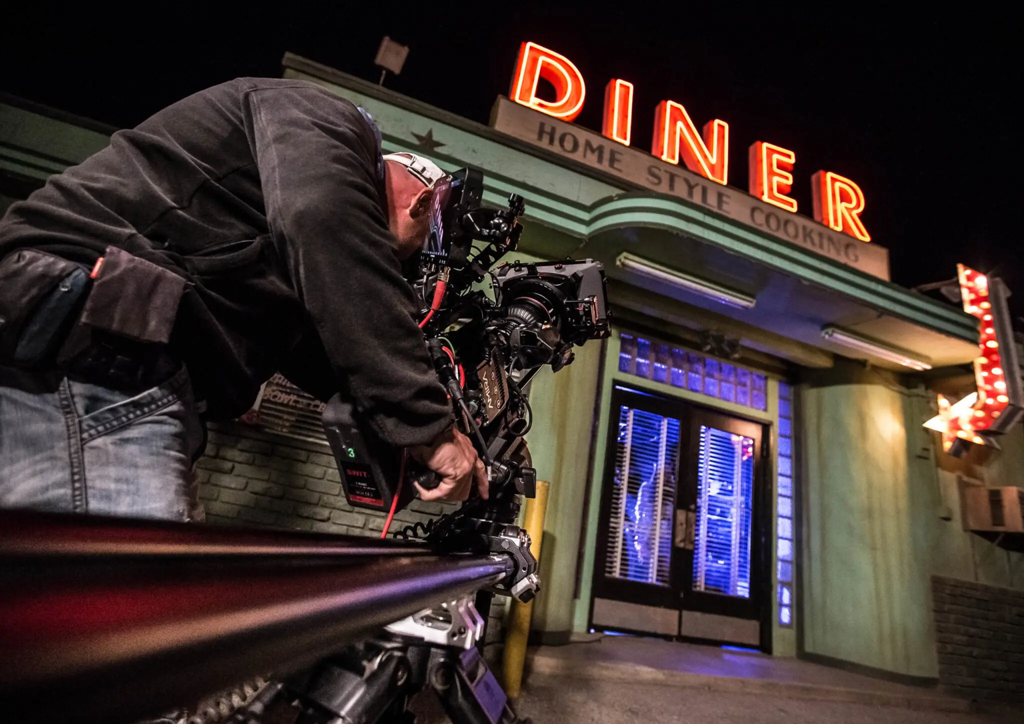 Cameraman Thomas filming outside a diner at night with a neon 'DINER' sign and blue-lit entrance doors.