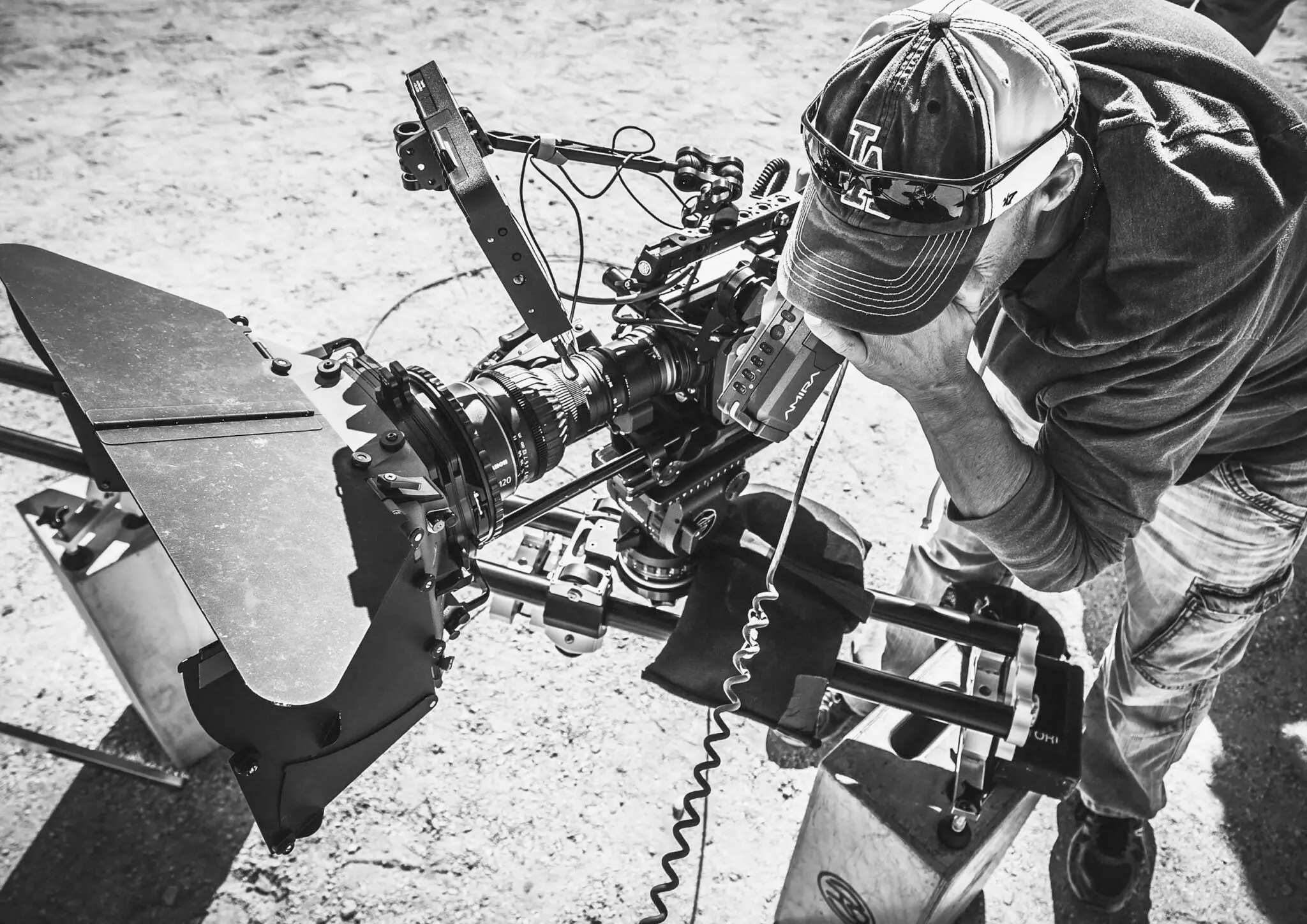 Man wearing a cap and sunglasses operating a professional camera setup with a matte box on a rig outdoors.