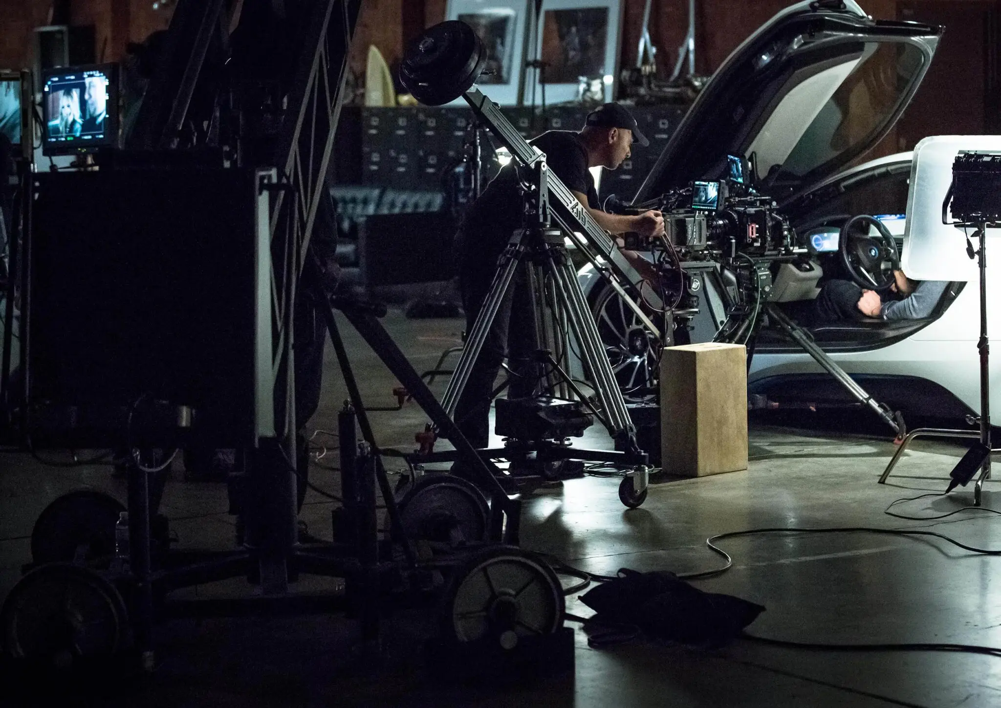 Crew filming inside a car with a camera crane and multiple monitors in a dimly lit studio.