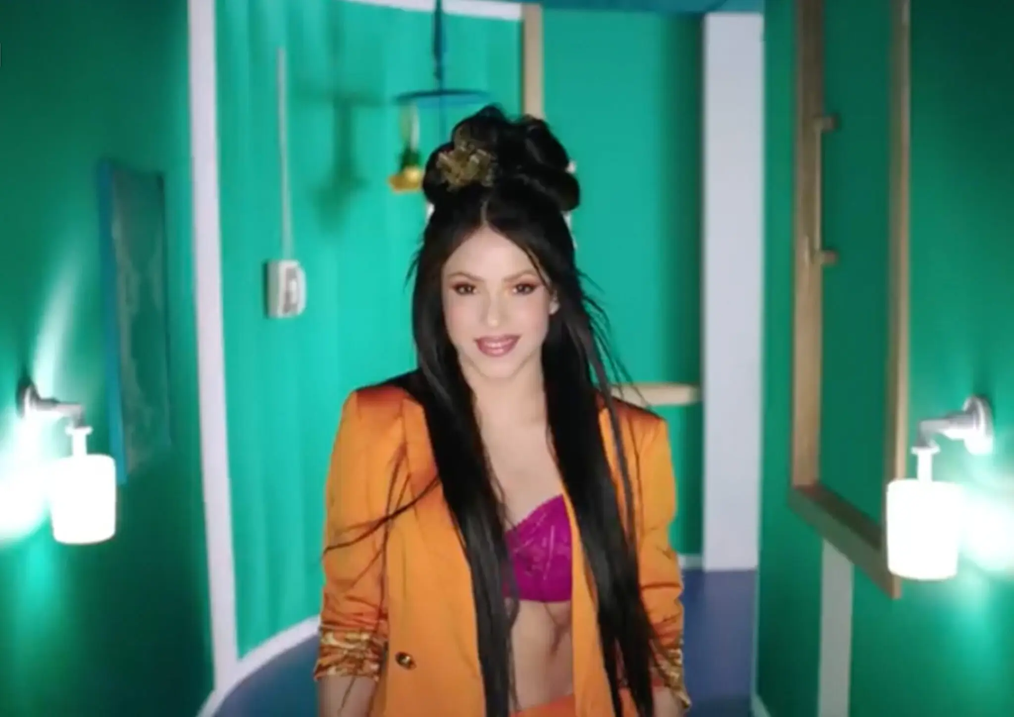 Smiling woman with long dark hair and an updo hairstyle wearing an orange jacket and purple cropped top in a teal hallway.