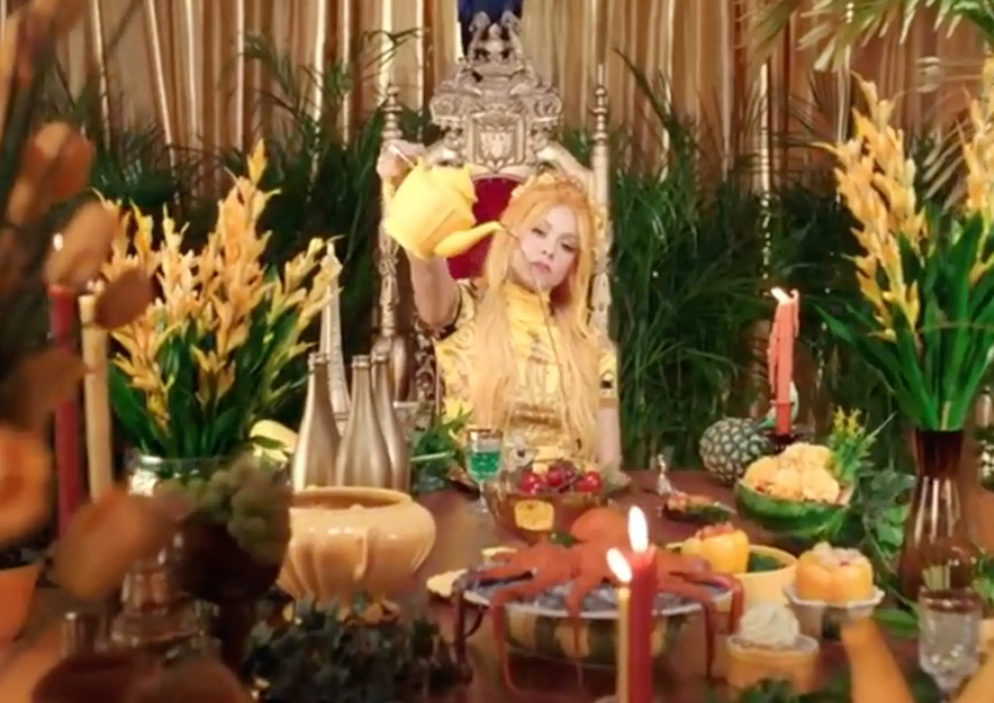 A woman in a yellow dress sits on an ornate throne pouring tea from a yellow teapot at a table filled with fruit, candles, and flowers.