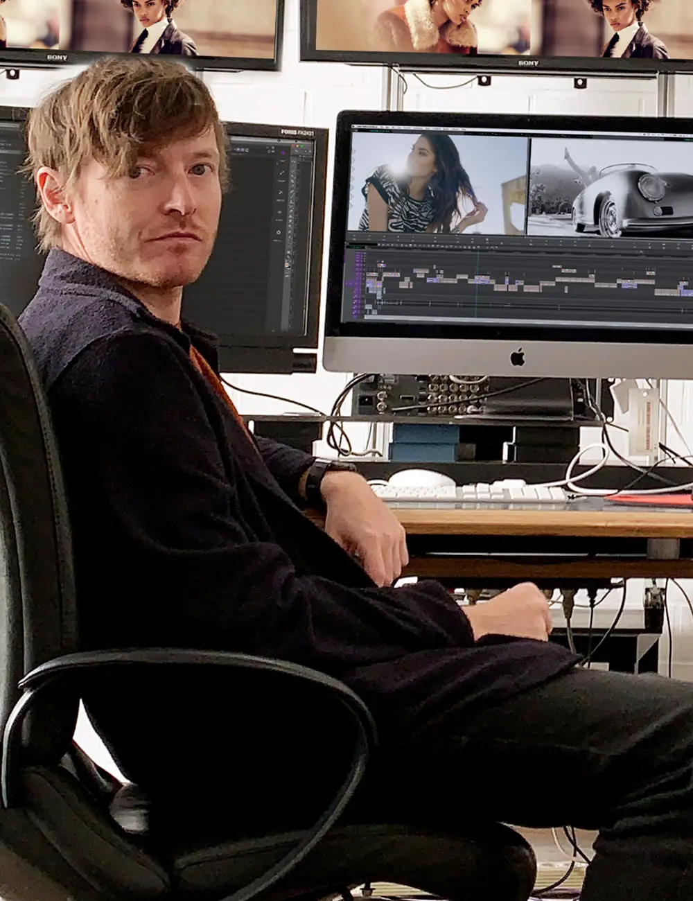 Ronald Rist sitting in an office chair at a desk with multiple monitors displaying video editing and photo software.
