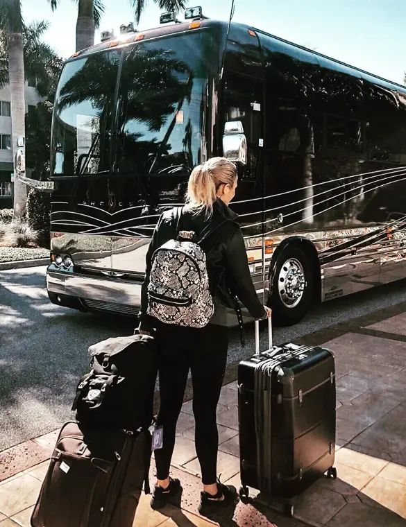Ferly with blonde ponytail wearing black clothes and a patterned backpack standing on a sidewalk next to a black tour bus, holding two rolling suitcases.