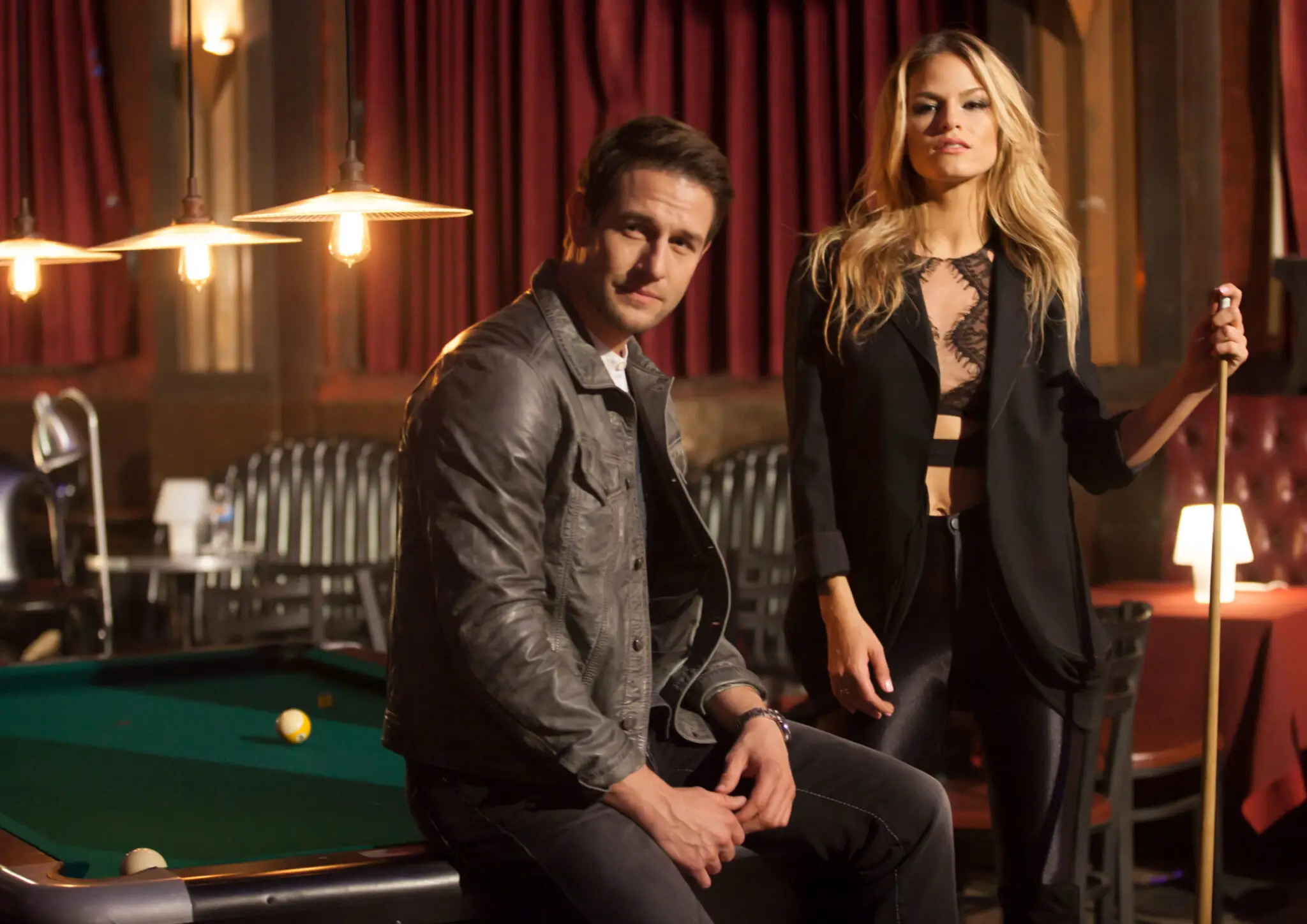 A man in a gray jacket is sitting on the edge of a pool table, and a Ferly Prado in black is holding a pool cue in a billiard room.