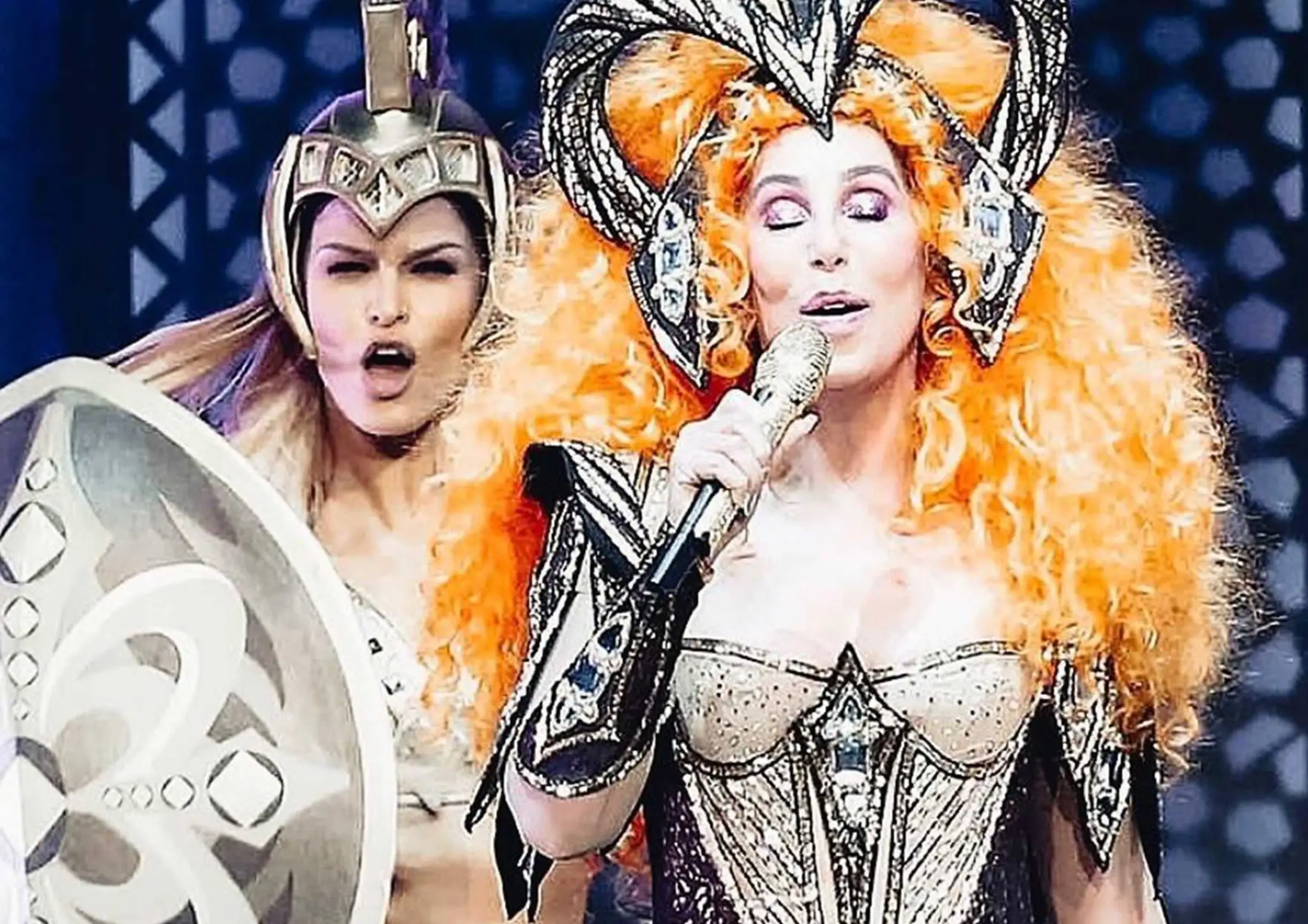 Performer with large curly orange hair and elaborate costume singing into a gold microphone, accompanied by a person in warrior helmet and shield.