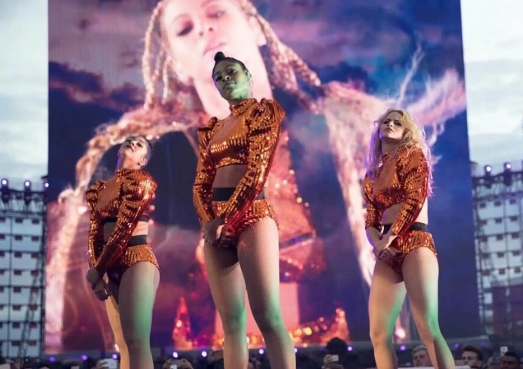 Three female dancers in matching orange sequin outfits performing on stage with a large video screen behind them.