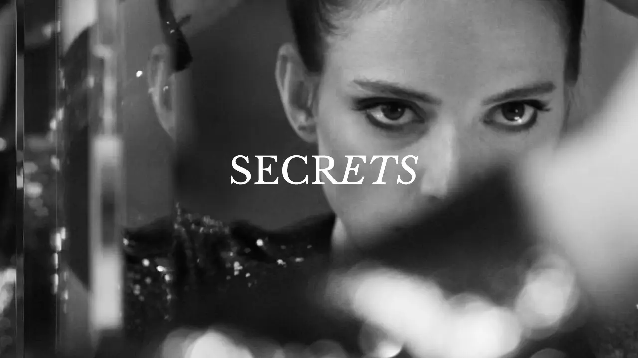 Black and white close-up of a woman looking into a mirror, with the word “SECRETS” overlaid in white text, shown in a screenshot from the movie Secrets.