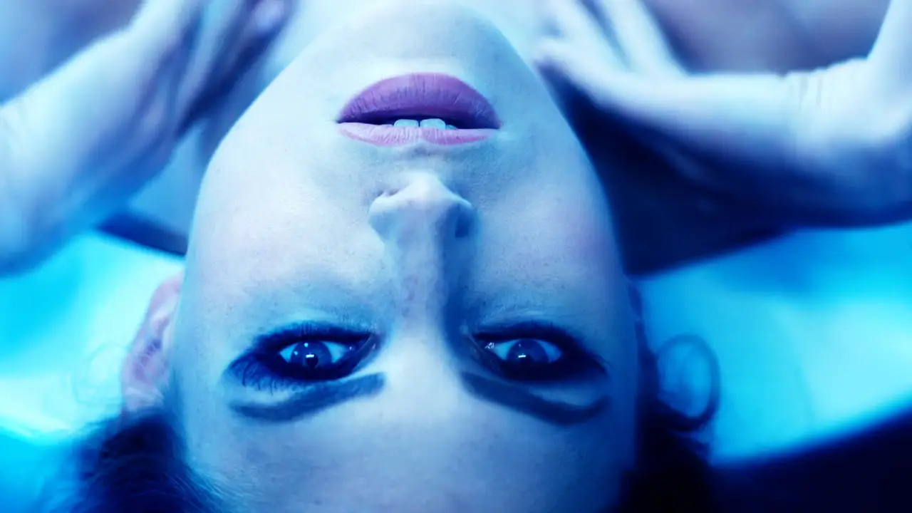 Screenshot from the movie Shadow of the Third showing a close-up of a woman’s face illuminated in blue light, lying down with her hands near her head and eyes wide open.