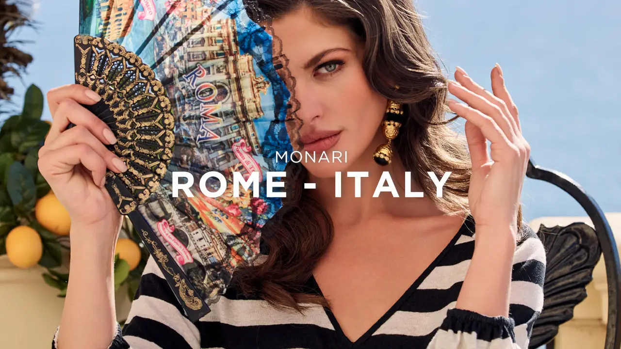 Woman with long dark hair holding a decorative fan featuring Rome landmarks, wearing gold earrings and a black-and-white striped top.