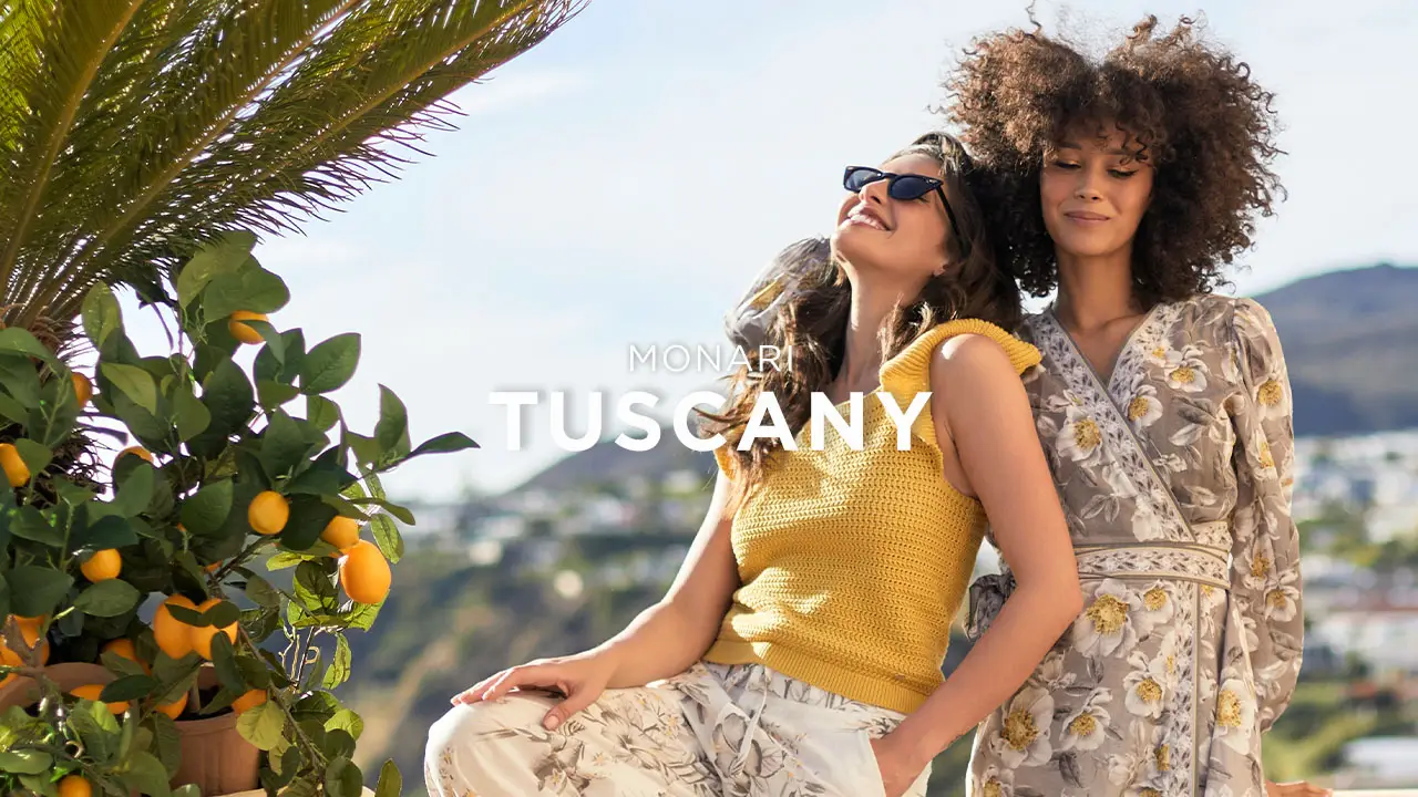 Two women in floral and yellow outfits posing outdoors near orange trees with hills in the background.