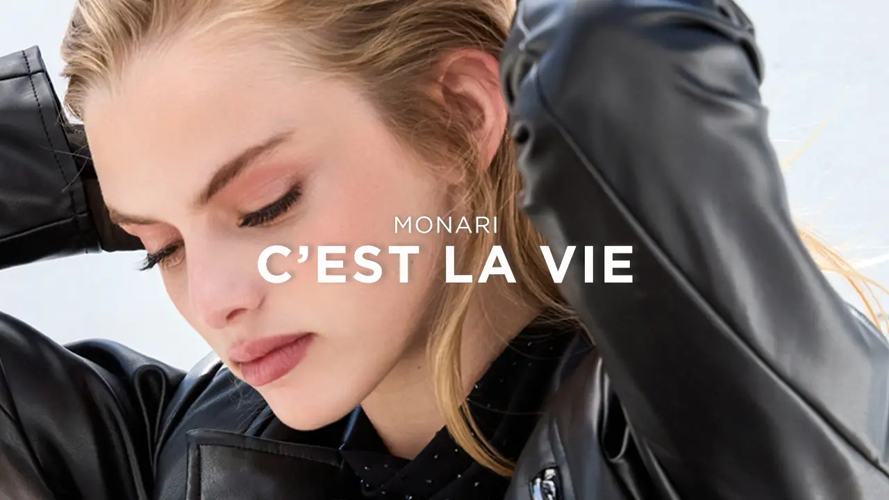 Close-up of a woman with blonde hair wearing a black leather jacket, looking down with her hands behind her head.