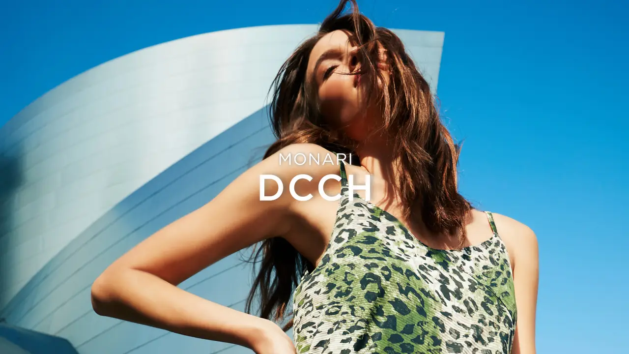 Woman with long hair in a green leopard print dress poses outdoors against a blue sky and modern building backdrop.