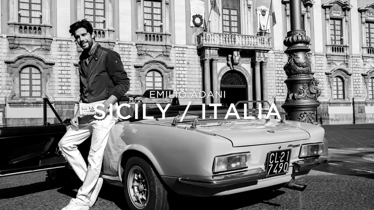 Man leaning against a vintage convertible car parked in front of a historic stone building in Sicily, Italy.