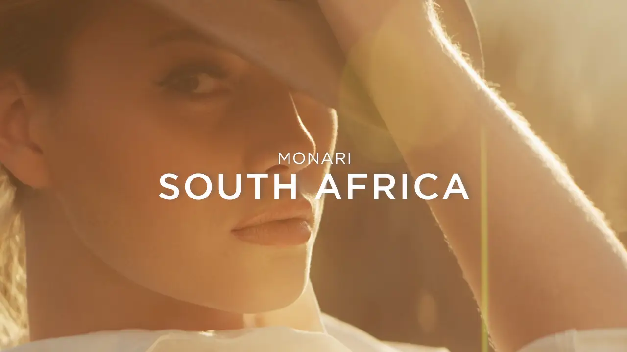Close-up of a woman wearing a hat, illuminated by warm sunlight, with text 'Monari South Africa' overlaid.