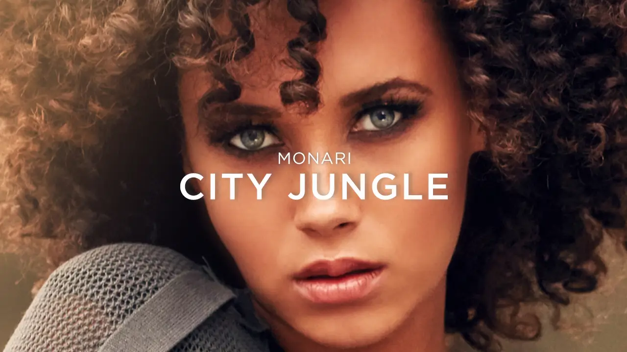 Close-up of a woman with curly hair, light eyes, and neutral makeup wearing a gray textured sweater.