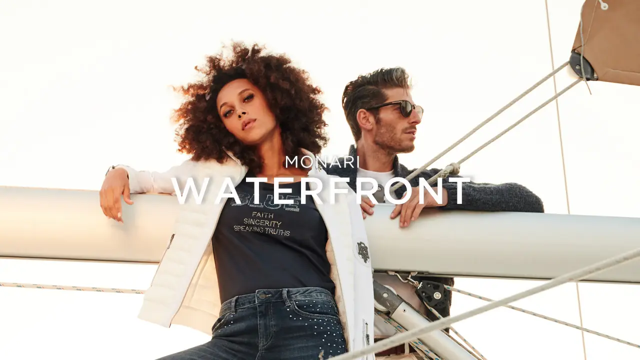 Fashionable woman with curly hair and man wearing sunglasses leaning on a sailboat mast with 'MONARI WATERFRONT' text overlay.