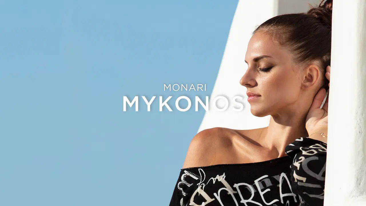 Woman with eyes closed leaning against a white wall wearing a black patterned top under a clear blue sky.