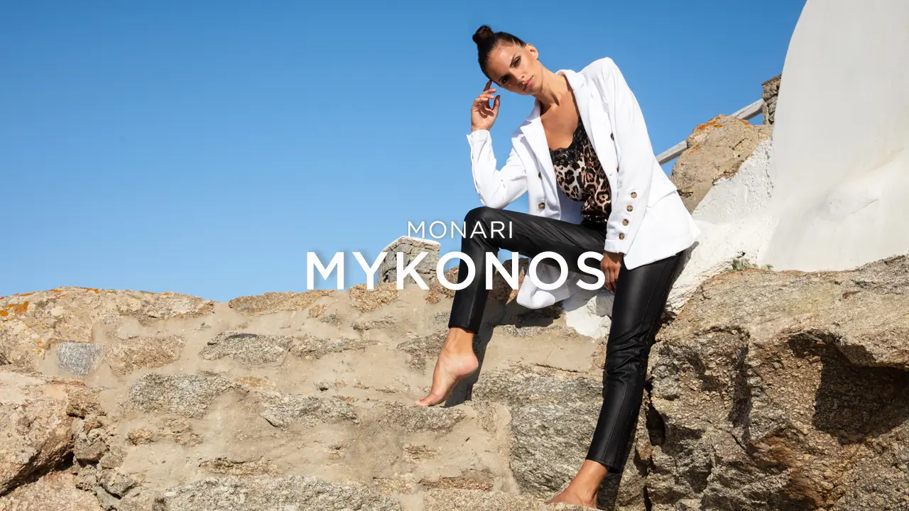 Woman in white blazer and black leather pants posing barefoot on stone steps under clear blue sky.