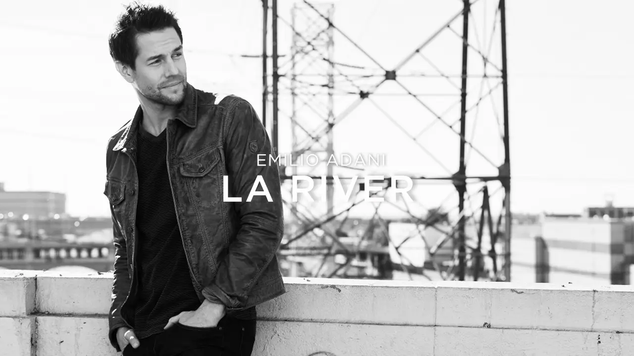 Man in denim jacket leaning on concrete ledge with urban industrial background and text 'Emilio Adani La River'.
