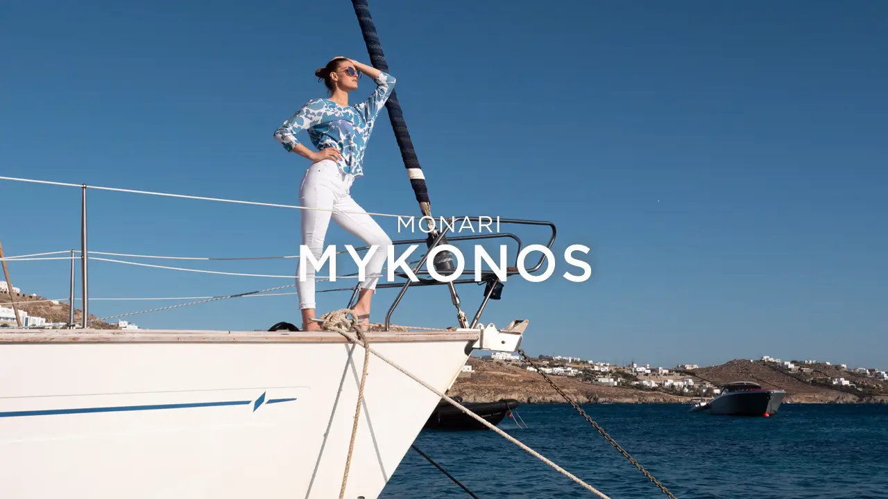 Woman in white pants and blue patterned shirt standing confidently on a boat bow overlooking the sea with a coastline in the background and clear blue sky.