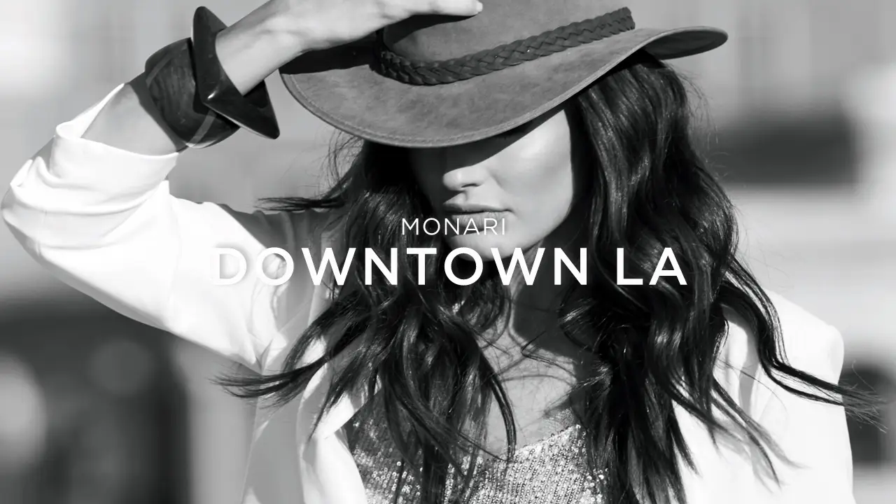 Black-and-white photo of a woman with long hair wearing a hat and white jacket, with text 'Monari Downtown LA' overlayed.