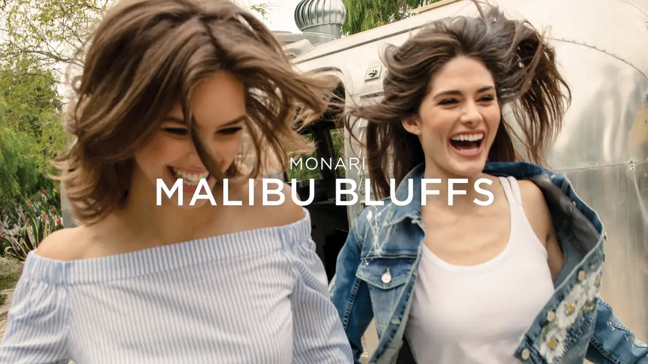 Two smiling young women with windblown hair, one in a striped off-shoulder top and the other in a white tank with a denim jacket, outdoors near a silver trailer.