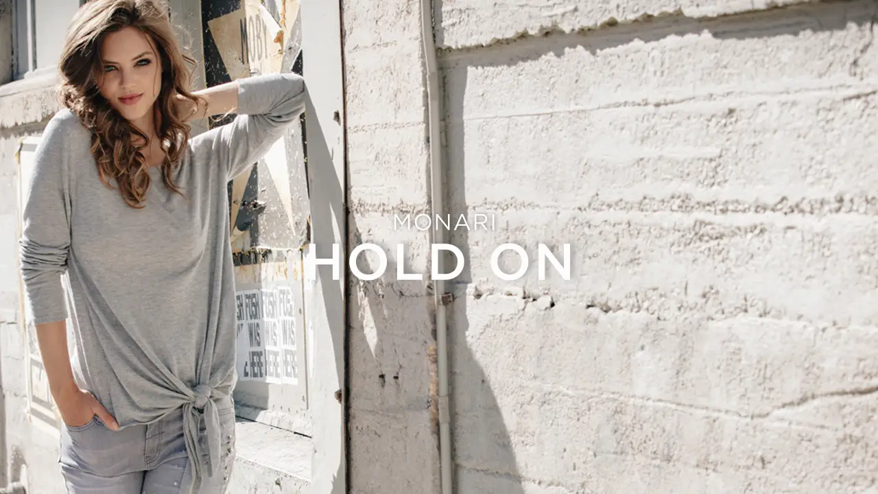 Woman in a light gray long-sleeve top with a front tie and gray pants standing against a textured white wall.