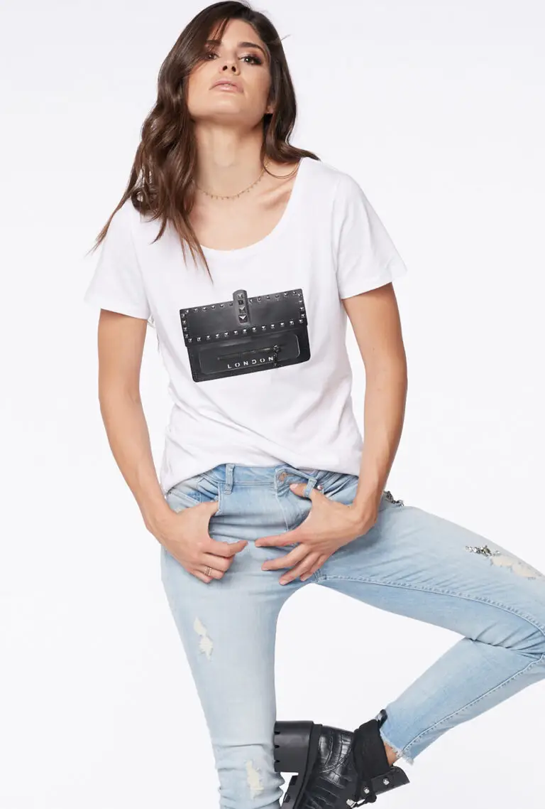 Woman with long brown hair wearing a white t-shirt with a black studded purse graphic labeled 'London', light ripped skinny jeans, and black ankle boots.