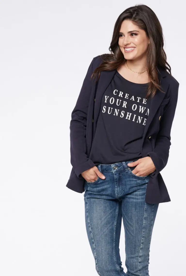 Smiling woman wearing navy blazer, black shirt with 'CREATE YOUR OWN SUNSHINE' text, and blue jeans.