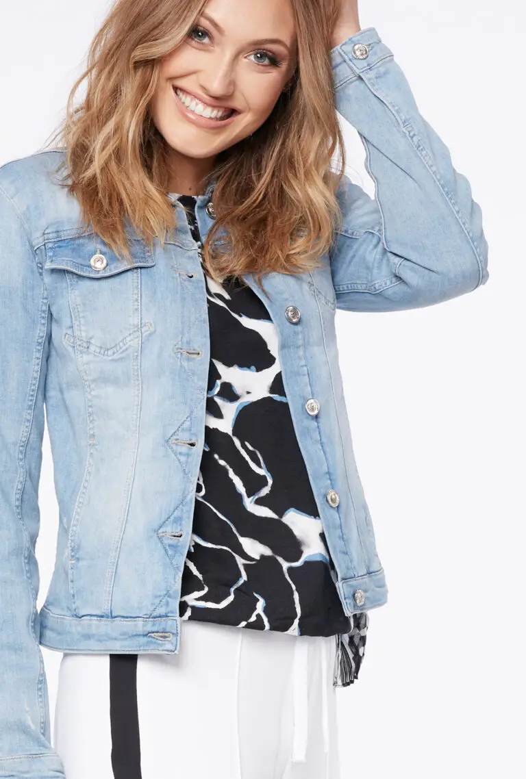 Smiling woman with shoulder-length blonde hair wearing a light blue denim jacket over a black and white patterned top.