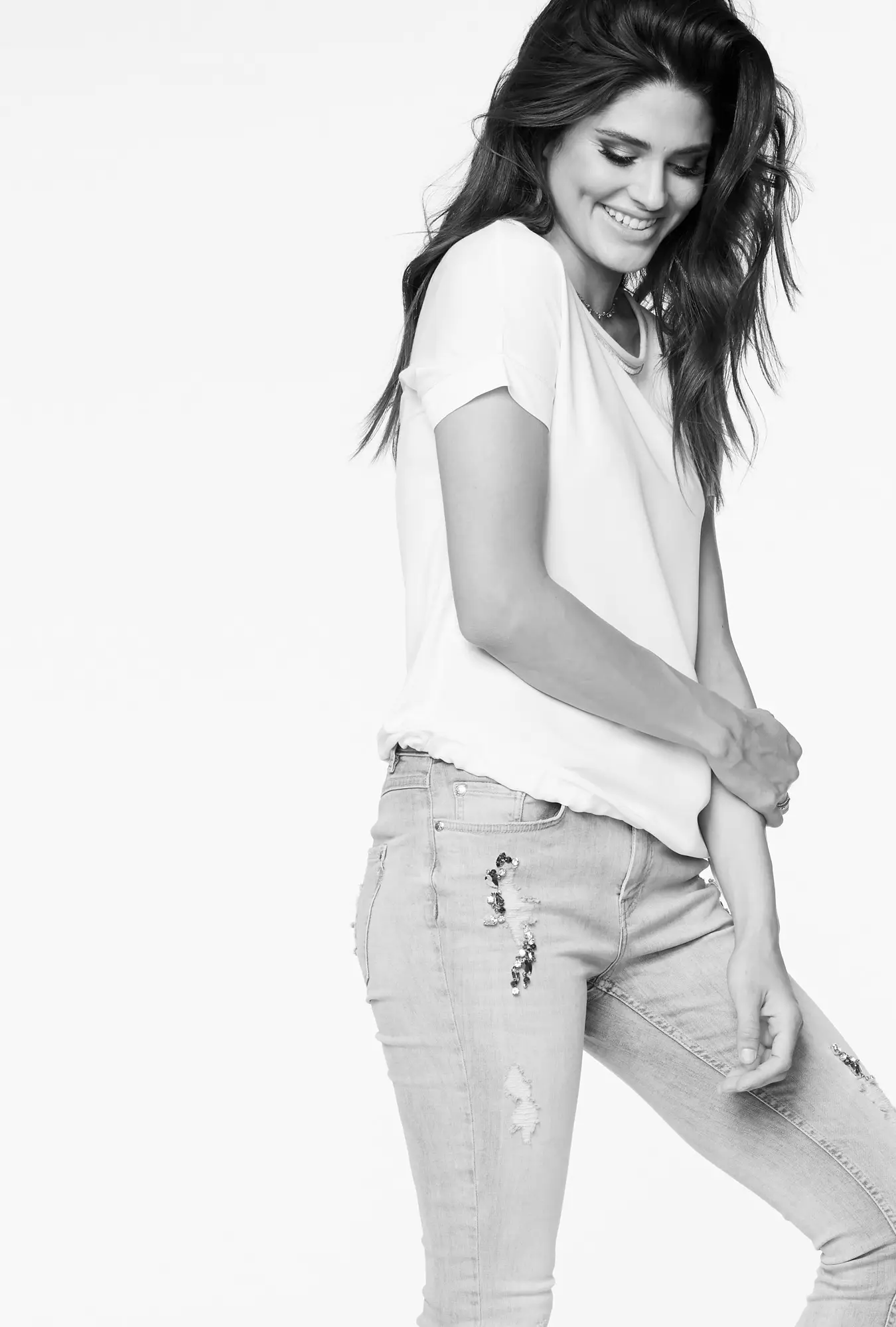 Smiling woman with long hair wearing a short-sleeve shirt and embellished ripped jeans, posing with arms crossed.