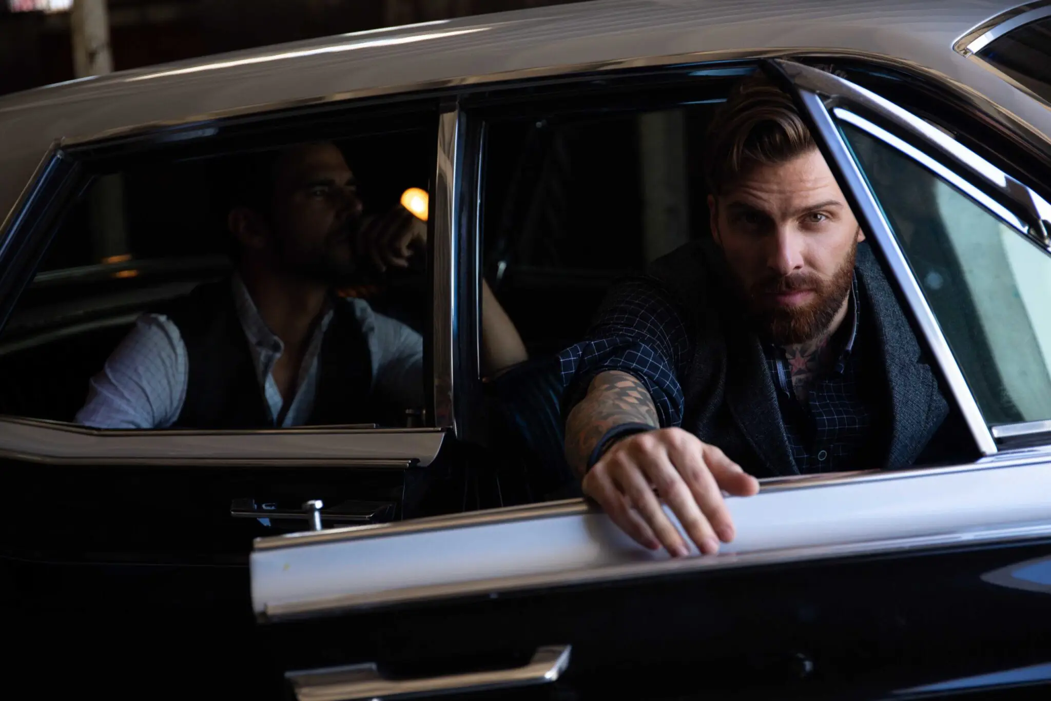 Bearded man with tattoos and a checked shirt opening the door of a black vintage car, with another man sitting inside in the background.