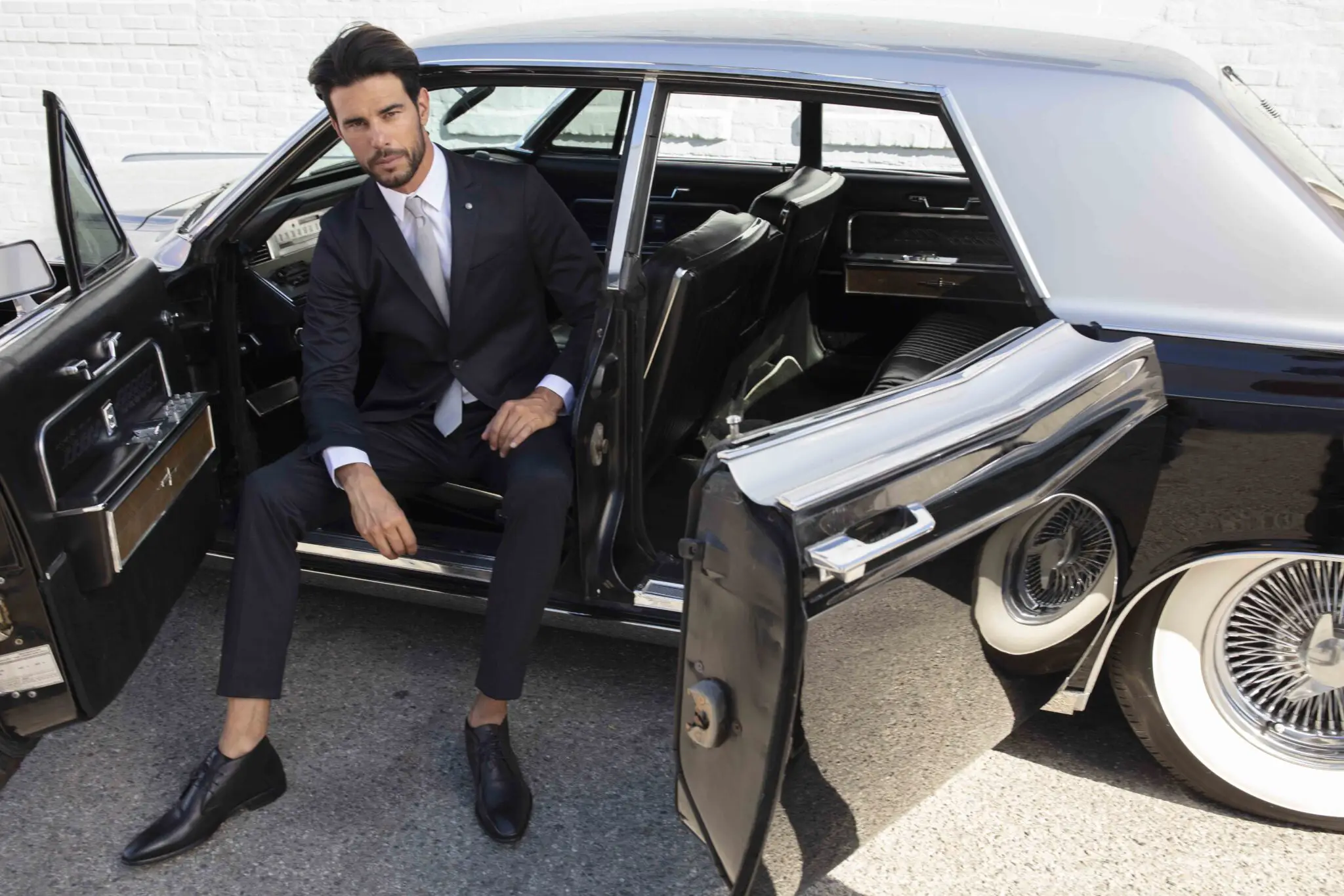Man in a dark suit and tie sitting on the open door sill of a vintage black car with whitewall tires.