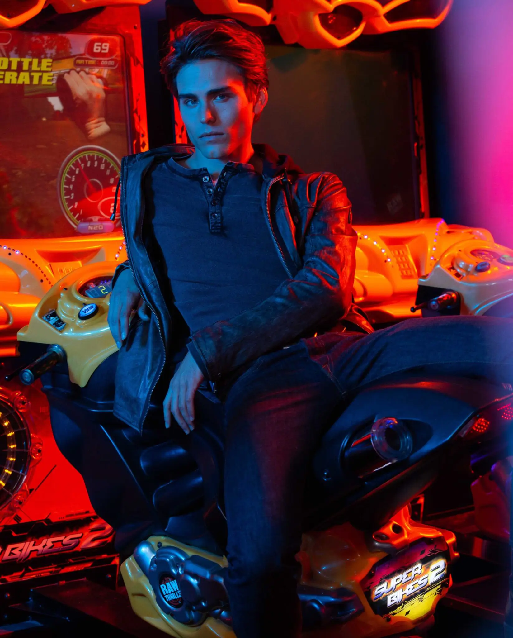 Young man with styled hair and leather jacket sitting on a yellow arcade motorcycle game in a room lit with red and blue lights.