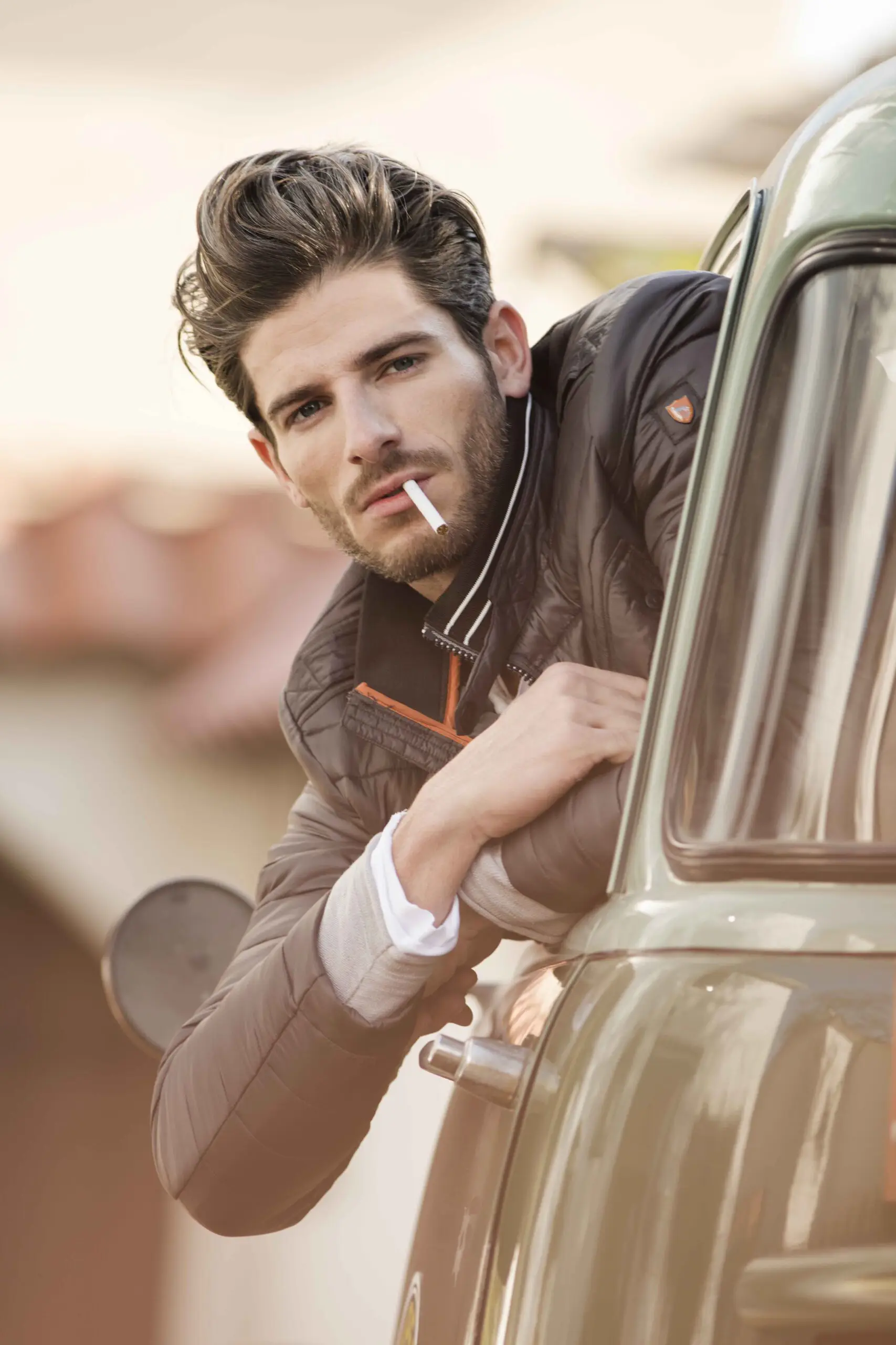 Man with styled hair and beard leaning out of a car window smoking a cigarette, wearing a dark jacket.