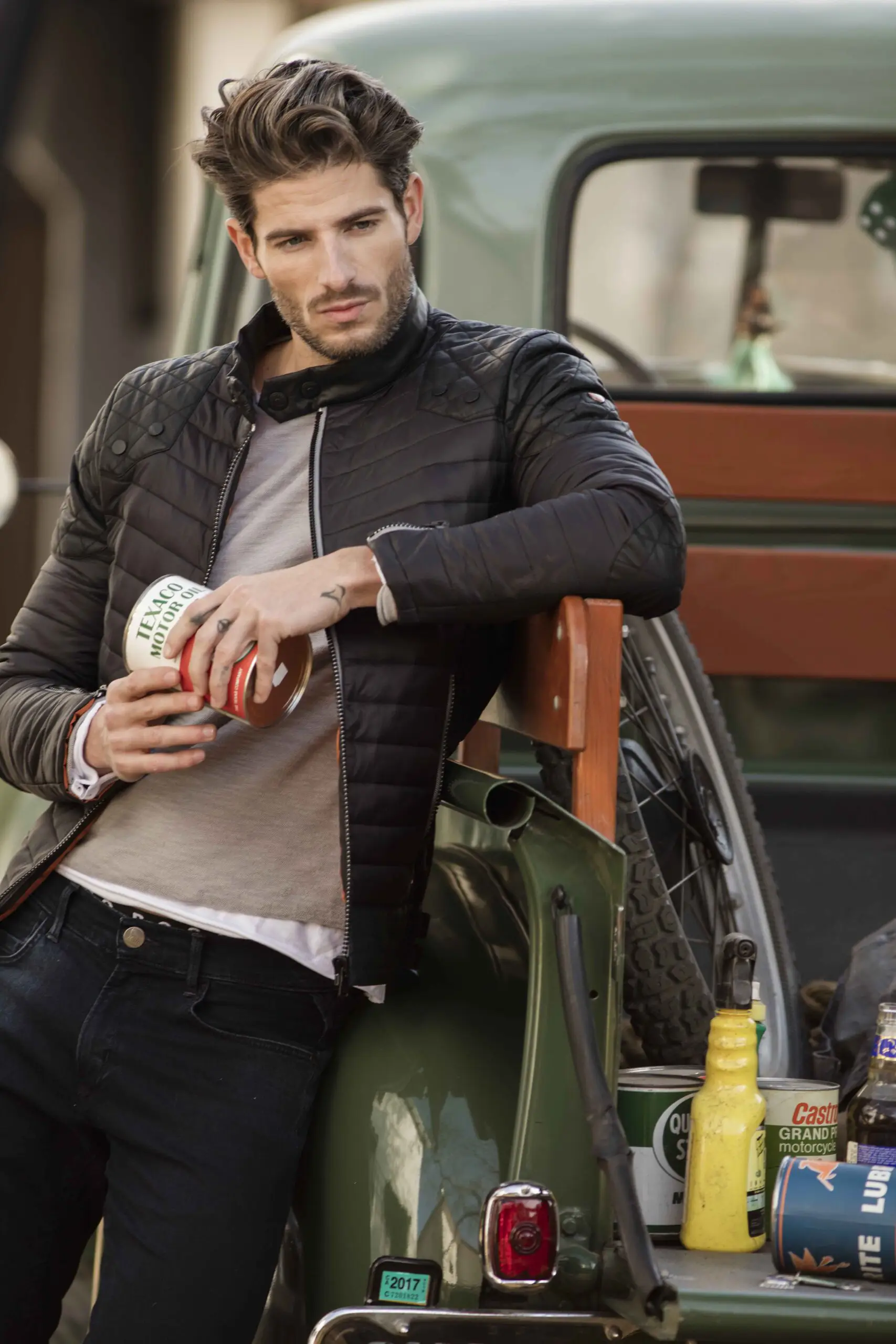 Man with styled hair in a black quilted jacket leaning against the back of a green pickup truck holding a Texaco motor oil can.