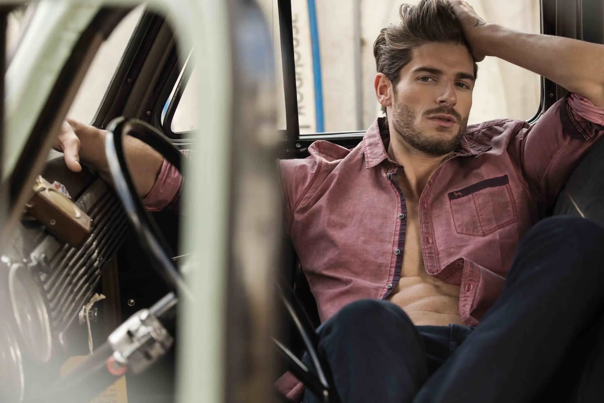 Man with beard lounging in a car seat, wearing a partially unbuttoned red shirt and black pants.