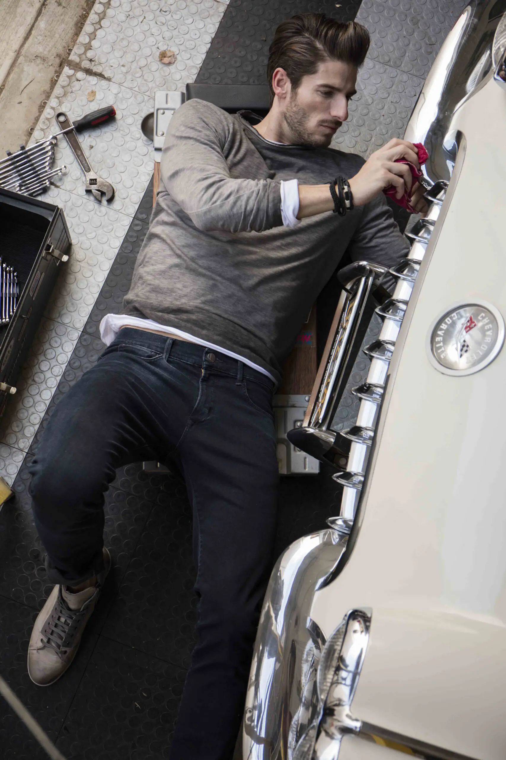Man lying on a mat working on a vintage white Chevrolet Corvette car, holding a red cloth near the front grille.