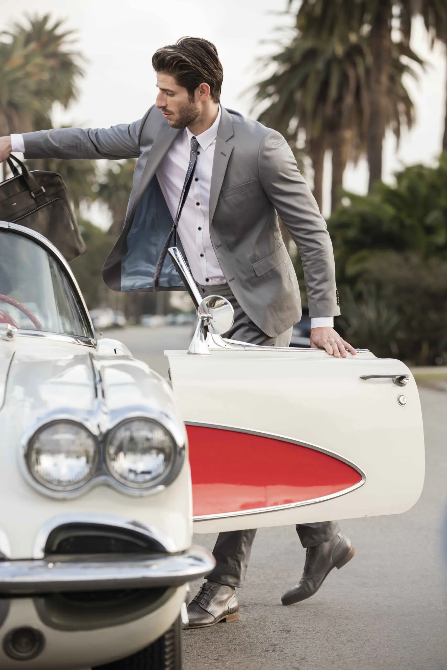 Man in gray suit opening the door of a white vintage car while holding a brown leather bag.