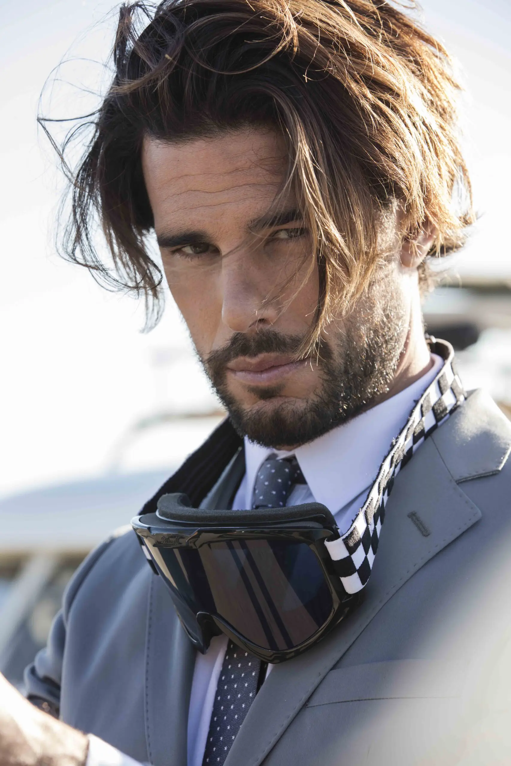 Close-up of a man with tousled hair and beard wearing a gray suit and tie with black goggles around his neck.