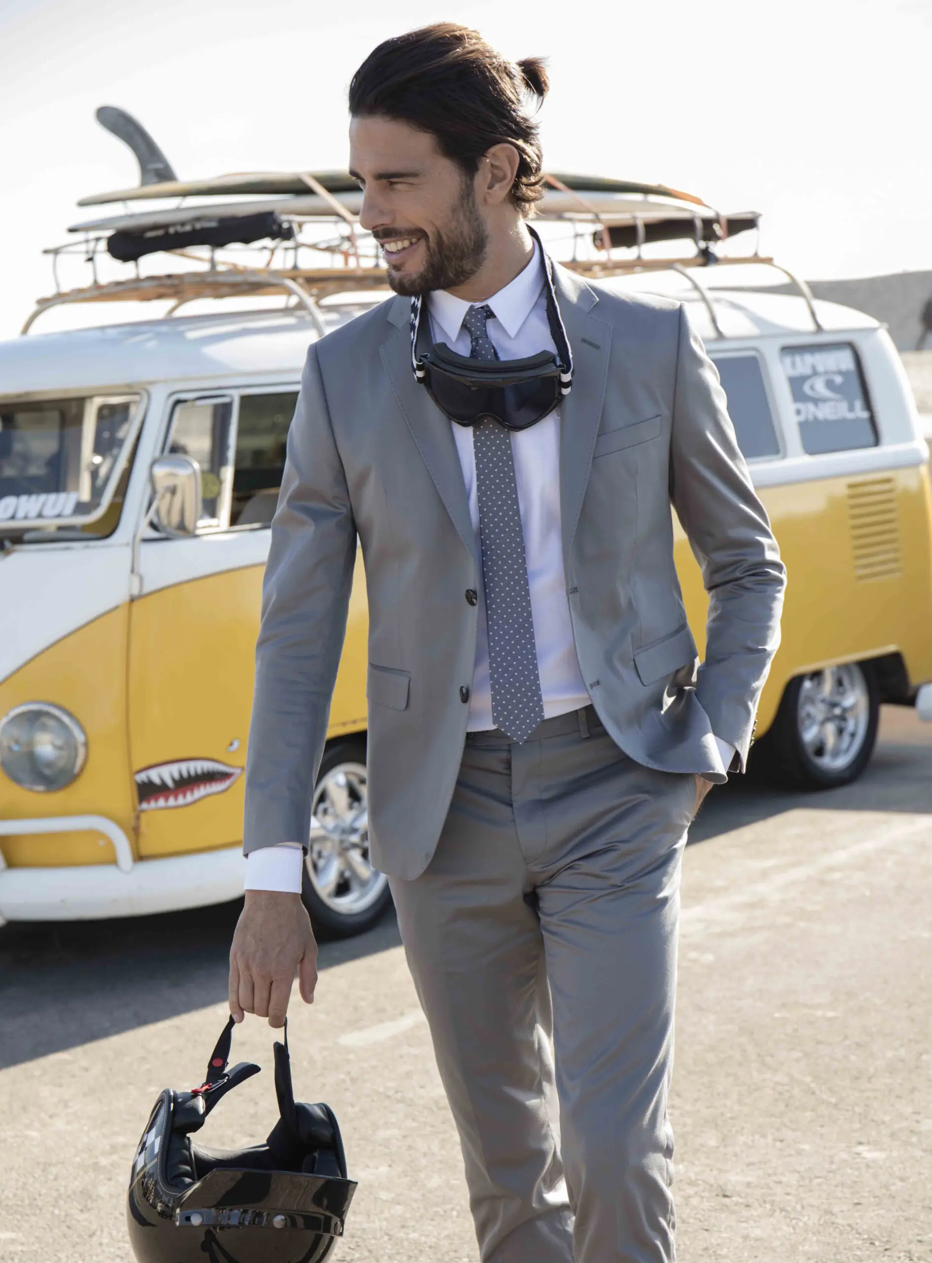 Man in a gray suit holding a black helmet, standing in front of a yellow and white vintage van with surfboards on the roof.