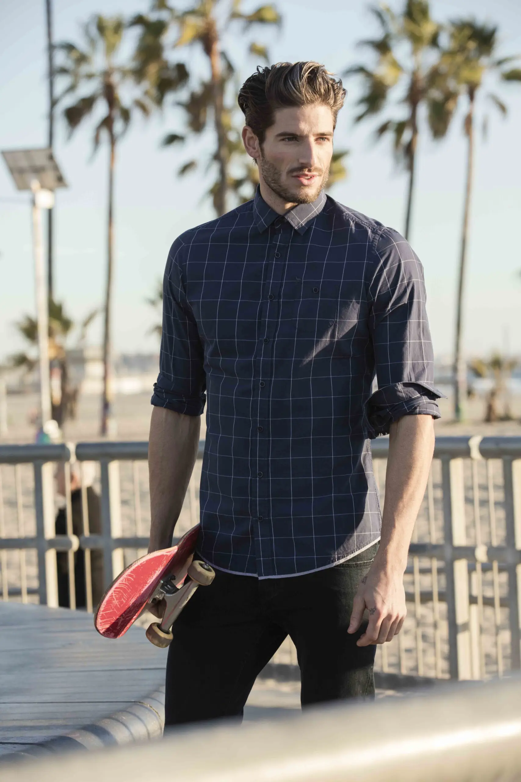 Man in a blue checkered shirt holding a red skateboard outdoors with palm trees in the background.