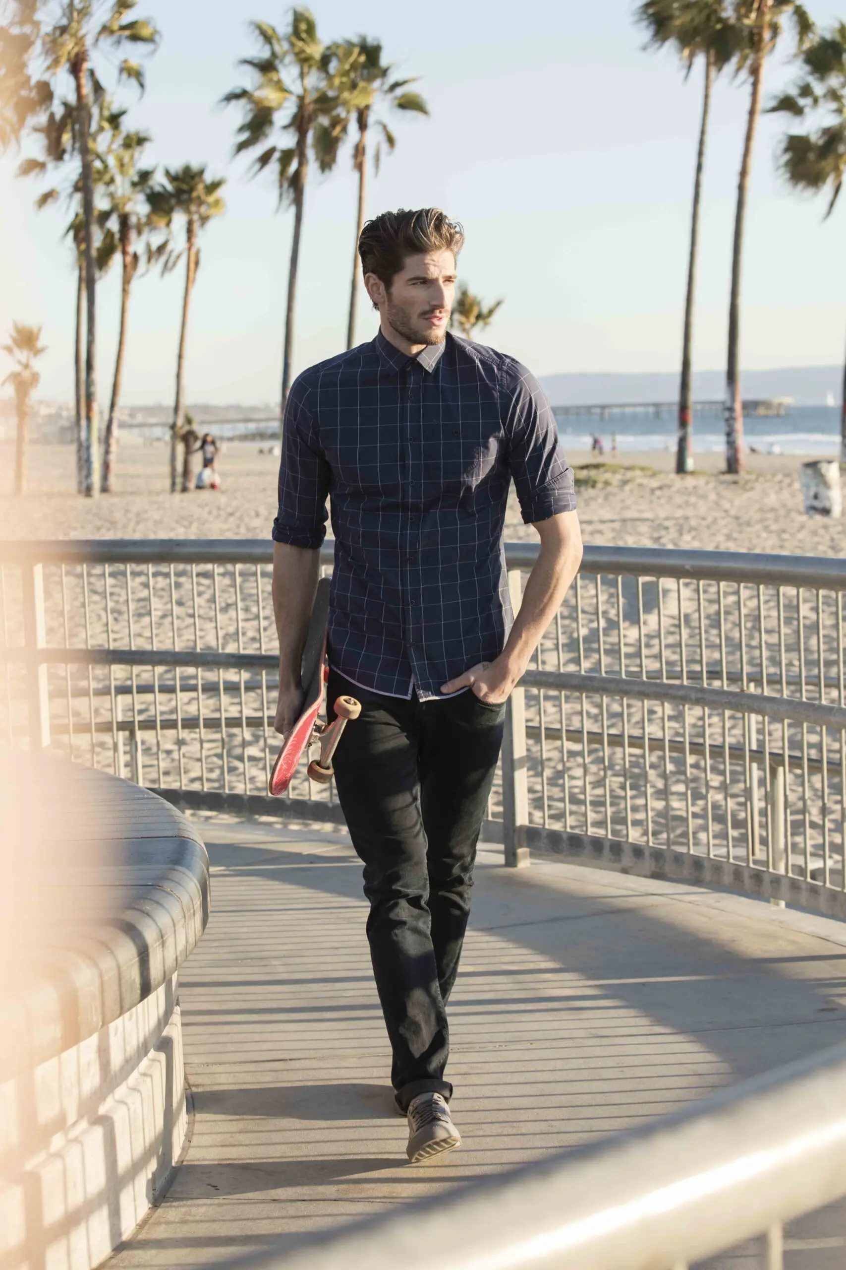 Man walking on beach promenade holding a skateboard, with palm trees and ocean in the background.